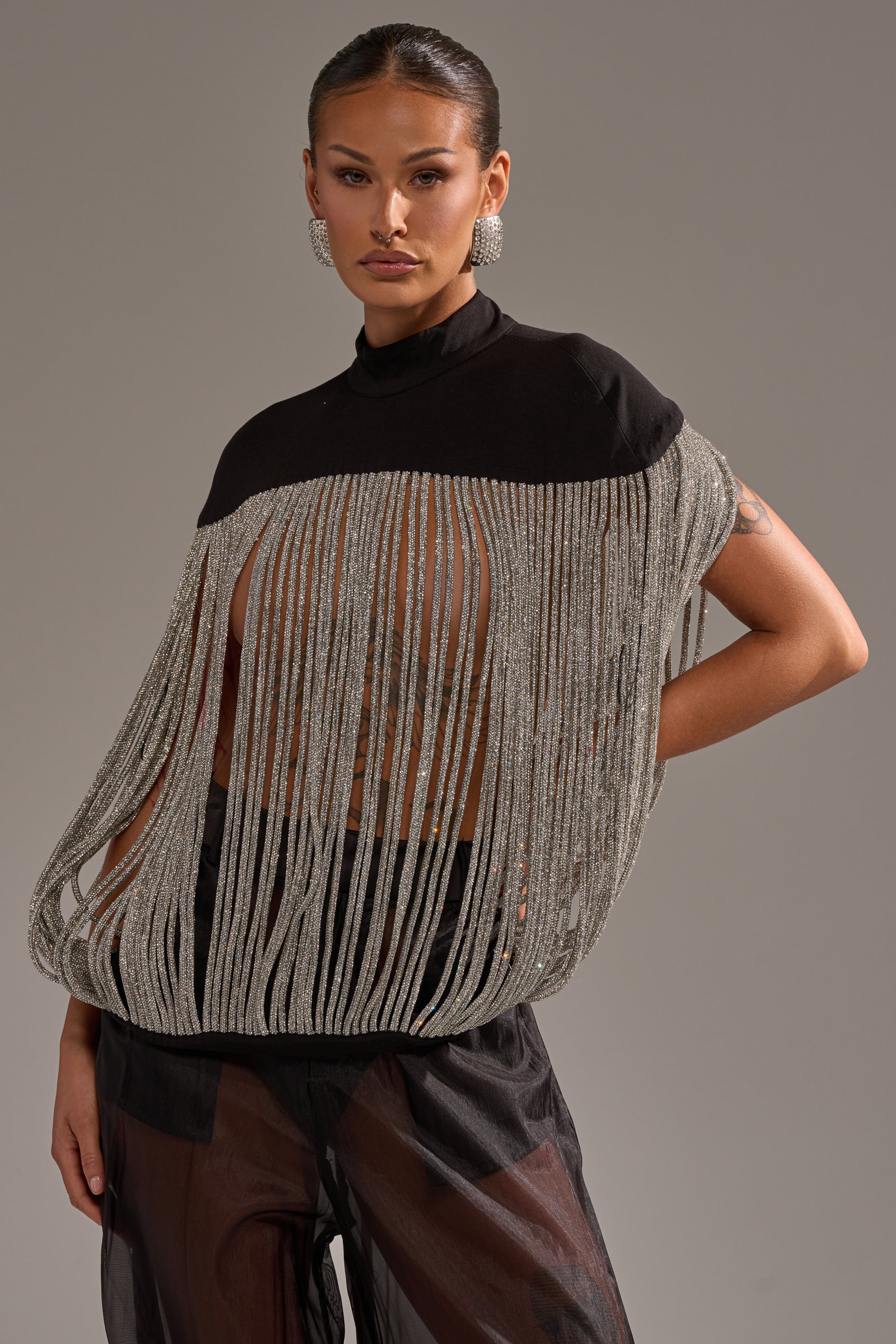 A woman models the DRIPPING IN DIAMONDS BLOUSE—a black top featuring long silver rhinestone fringe that reveals her torso—styled with sheer black pants and bold earrings, her slicked-back hair set against a plain gray backdrop.
