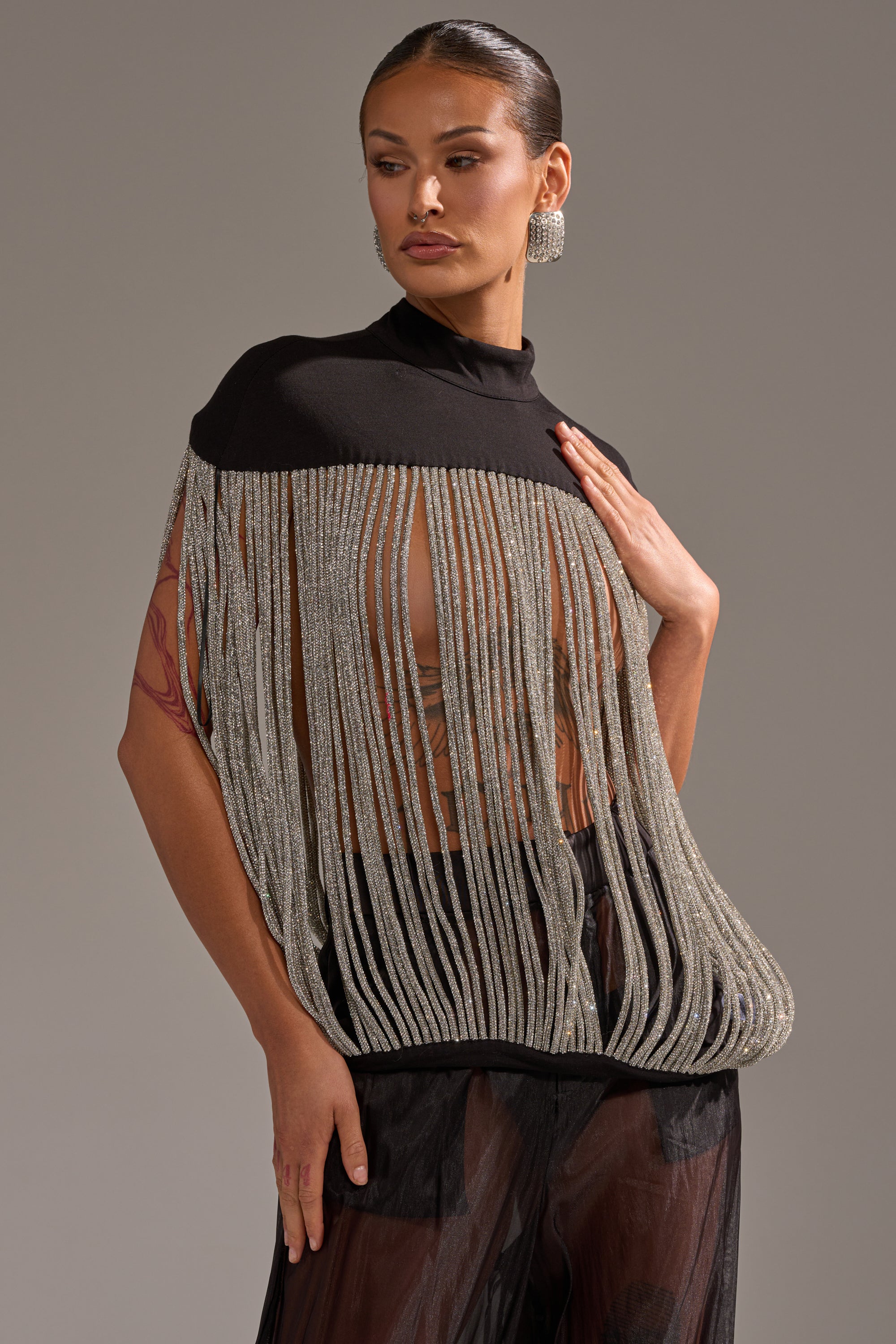 A woman wears the DRIPPING IN DIAMONDS BLOUSE—a sheer black top with rhinestone fringe and a high neckline—paired with sheer black pants. Her slicked-back hair, bold earrings, and confident demeanor complete the look.