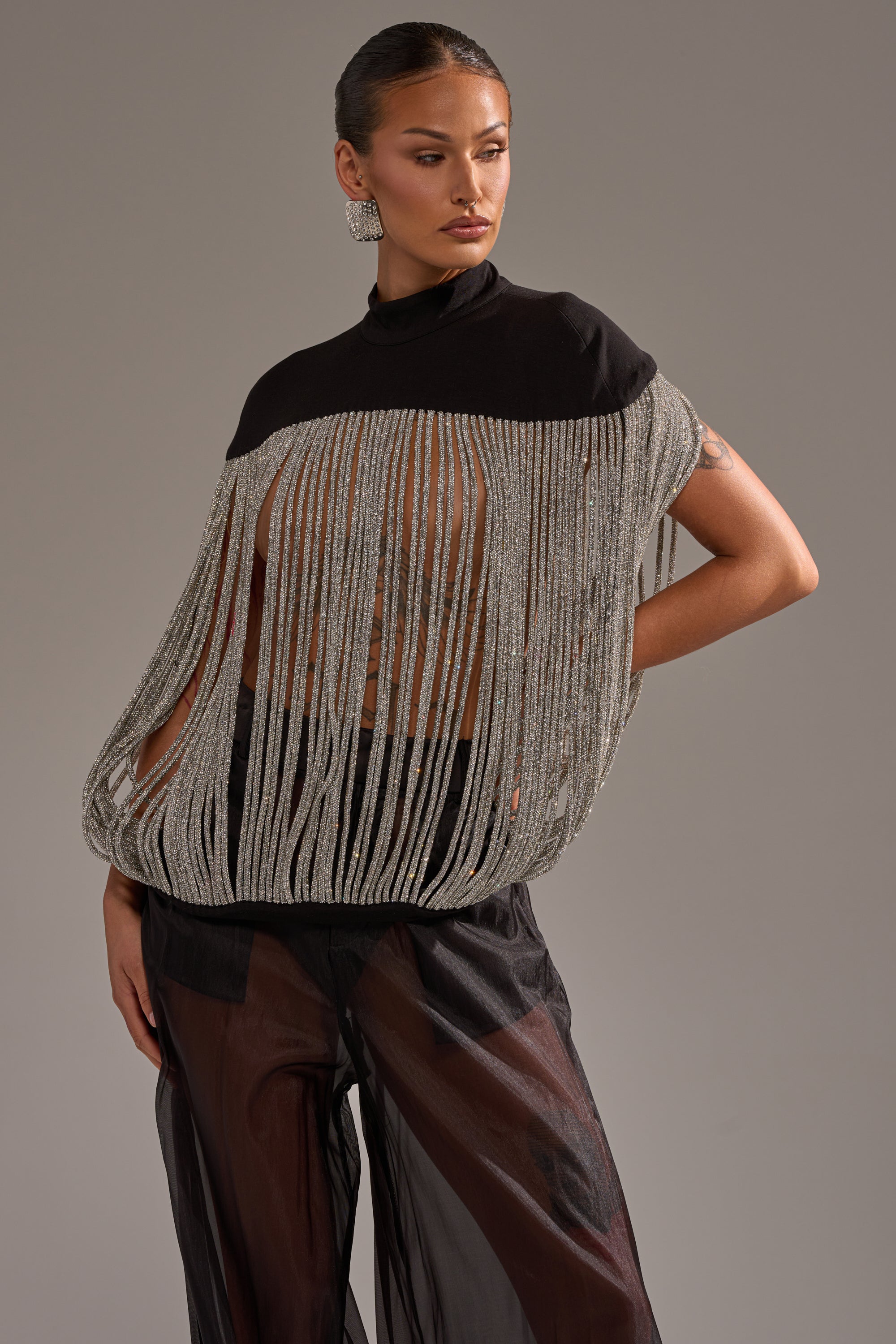 A woman poses confidently against a plain gray background in the DRIPPING IN DIAMONDS BLOUSE, styled with sheer black wide-leg pants. Statement earrings and slicked-back hair complete her striking look.