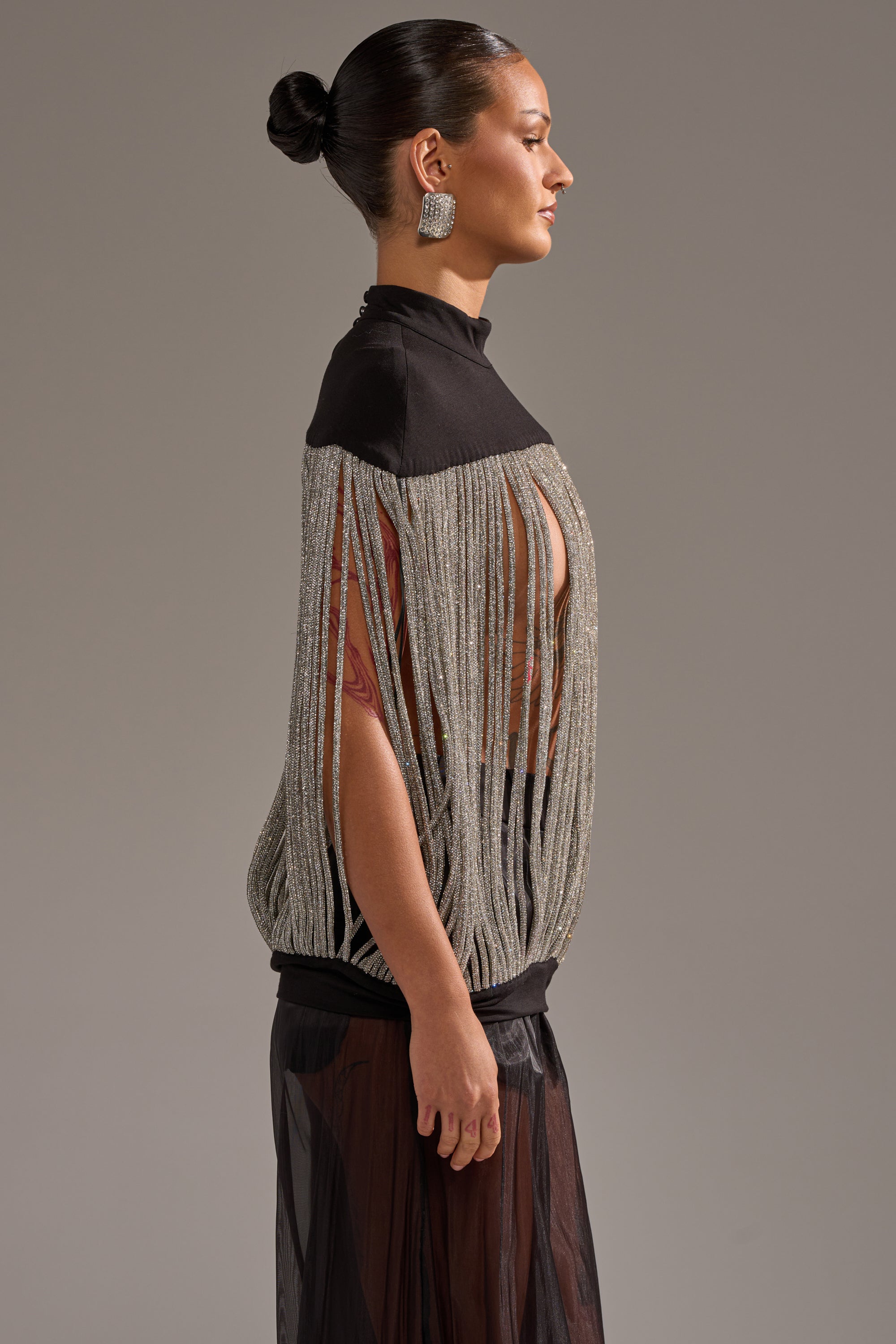A woman stands in profile against a plain background, wearing the DRIPPING IN DIAMONDS BLOUSE, a rhinestone fringe top with sheer black fabric at the bottom. Her hair is slicked back into a bun, paired with large square earrings.
