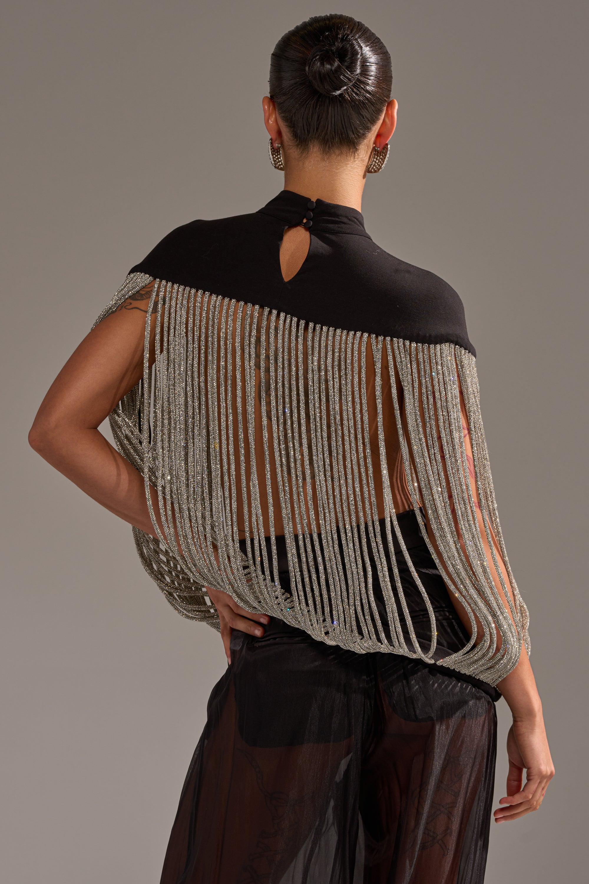 A person with hair in a bun models the DRIPPING IN DIAMONDS BLOUSE, featuring rhinestone fringe and long, draped silver chains across the back, paired with sheer black pants against a plain background.