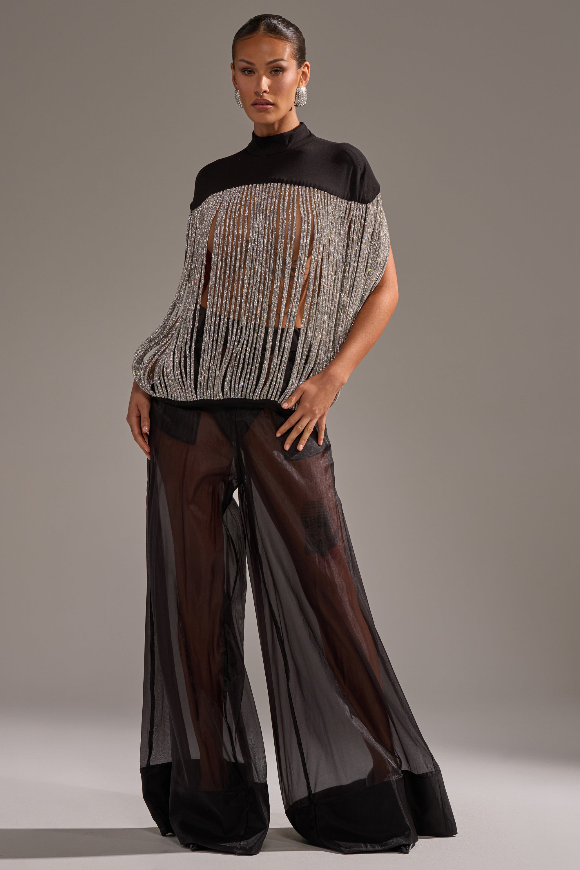 A woman poses in the sheer, loose DRIPPING IN DIAMONDS BLOUSE with metallic fringe over a black bandeau and wide-leg, see-through black pants, set against a plain light gray background.