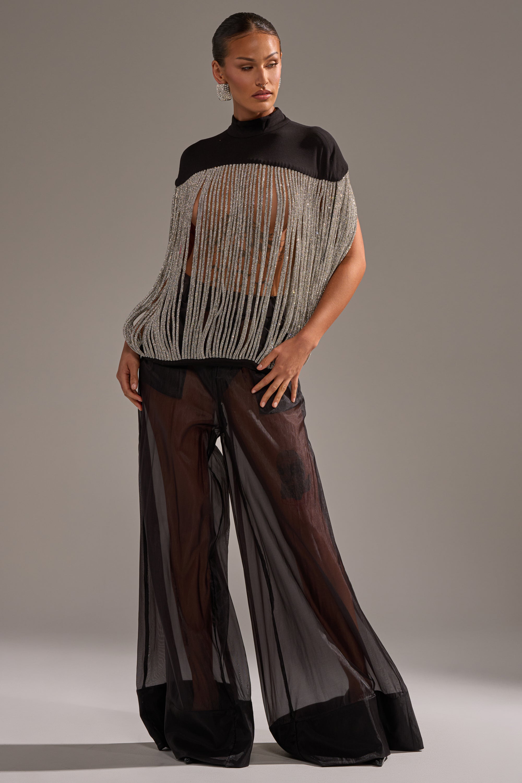 A woman stands confidently against a neutral background, wearing the DRIPPING IN DIAMONDS BLOUSE— a sheer black top with rhinestone fringe— paired with high-waisted, wide-leg black sheer pants over black underwear.