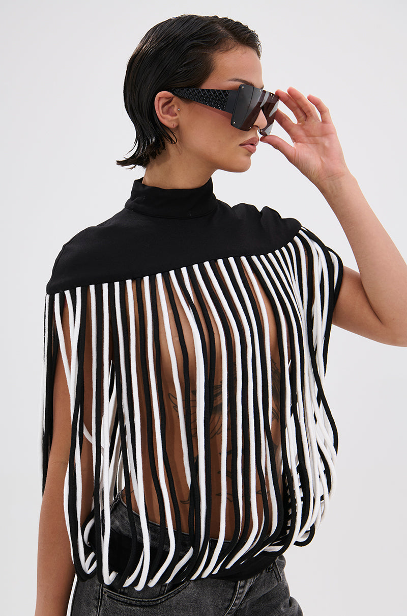 A person with short, wet-look hair and large black sunglasses confidently poses against a plain background, wearing the POWER TRIP FRINGE BLOUSE—a bold top with long black and white fringe—paired with dark jeans that reveal chest tattoos.