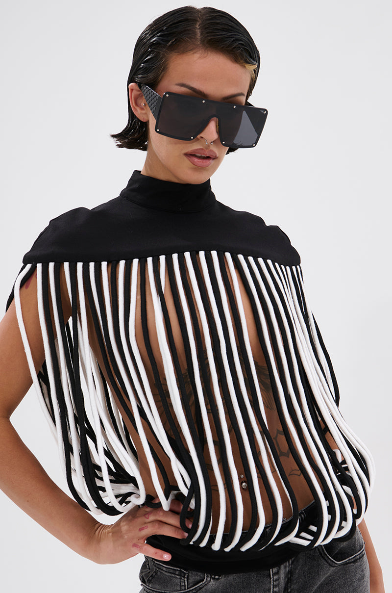 A person with short dark hair and square sunglasses wears the POWER TRIP FRINGE BLOUSE, featuring bold vertical black and white fringe strips and a sculptural mock neckline, revealing torso tattoos, styled with black jeans.