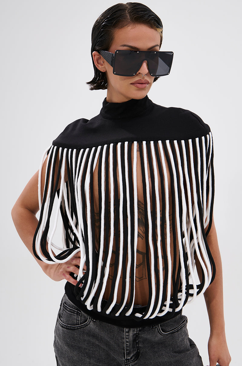 A person in oversized square sunglasses, dark jeans, and the POWER TRIP FRINGE BLOUSE with a sculptural mock neckline confidently poses with one hand on their hip against a plain background.