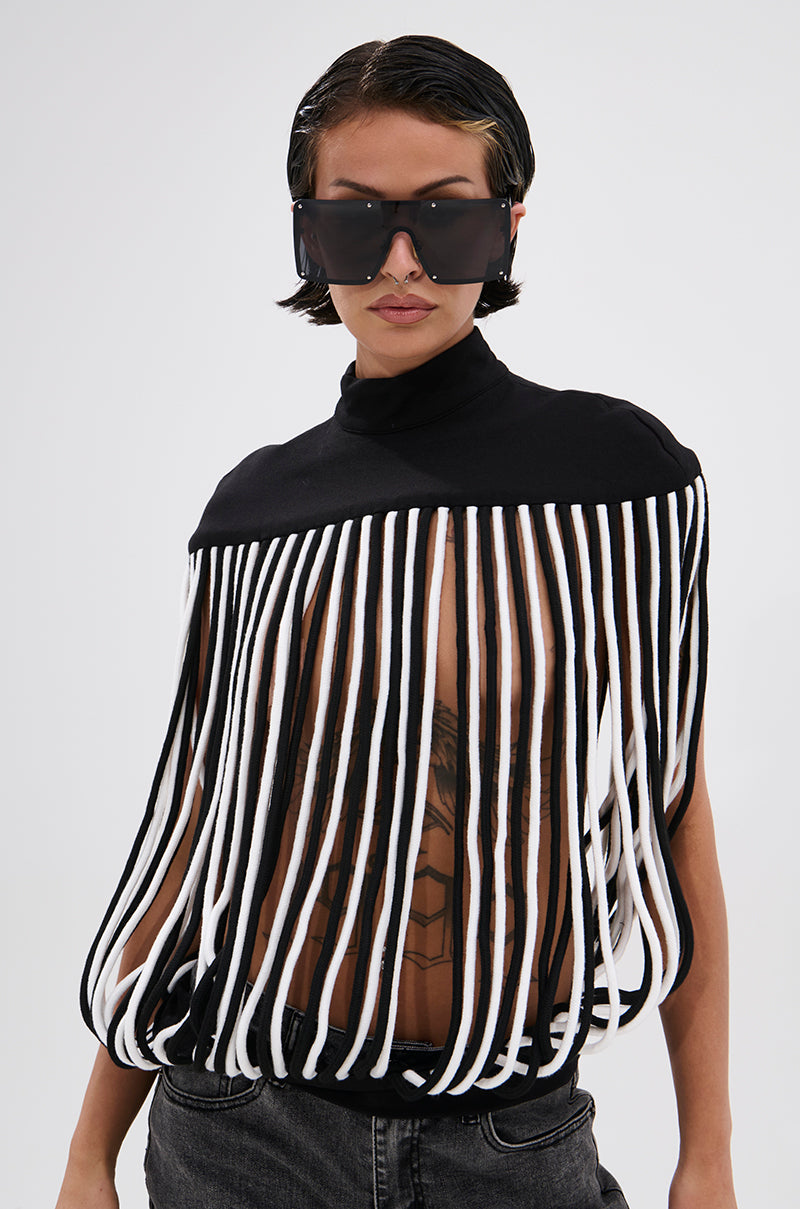 A person with slicked-back hair wears oversized black sunglasses and the POWER TRIP FRINGE BLOUSE—this striking top has a sculptural mock neckline and bold vertical black and white striped fringe, revealing tattoos beneath, set against a plain white background.