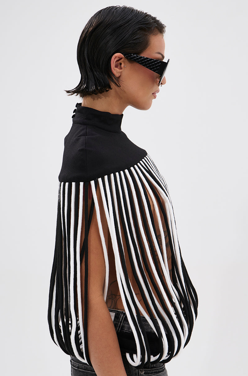 Wearing the POWER TRIP FRINGE BLOUSE—featuring a sculptural mock neck and bold black-and-white vertical fringe strips—a person with short, wet-look hair and sunglasses poses against a plain, light background, arms and sides exposed.