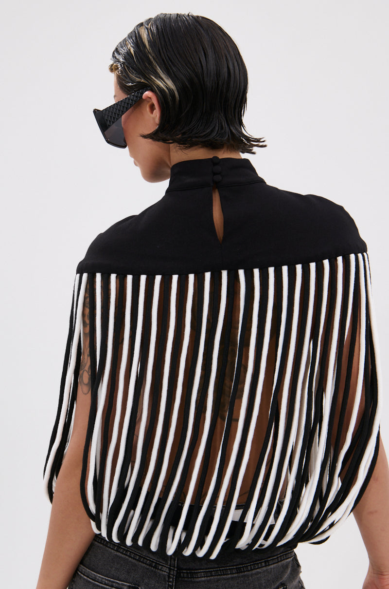 A person with slicked-back hair and sunglasses is seen from behind in the POWER TRIP FRINGE BLOUSE, featuring a sculptural mock neckline, long black-and-white fringe, and an open back that reveals a tattoo.