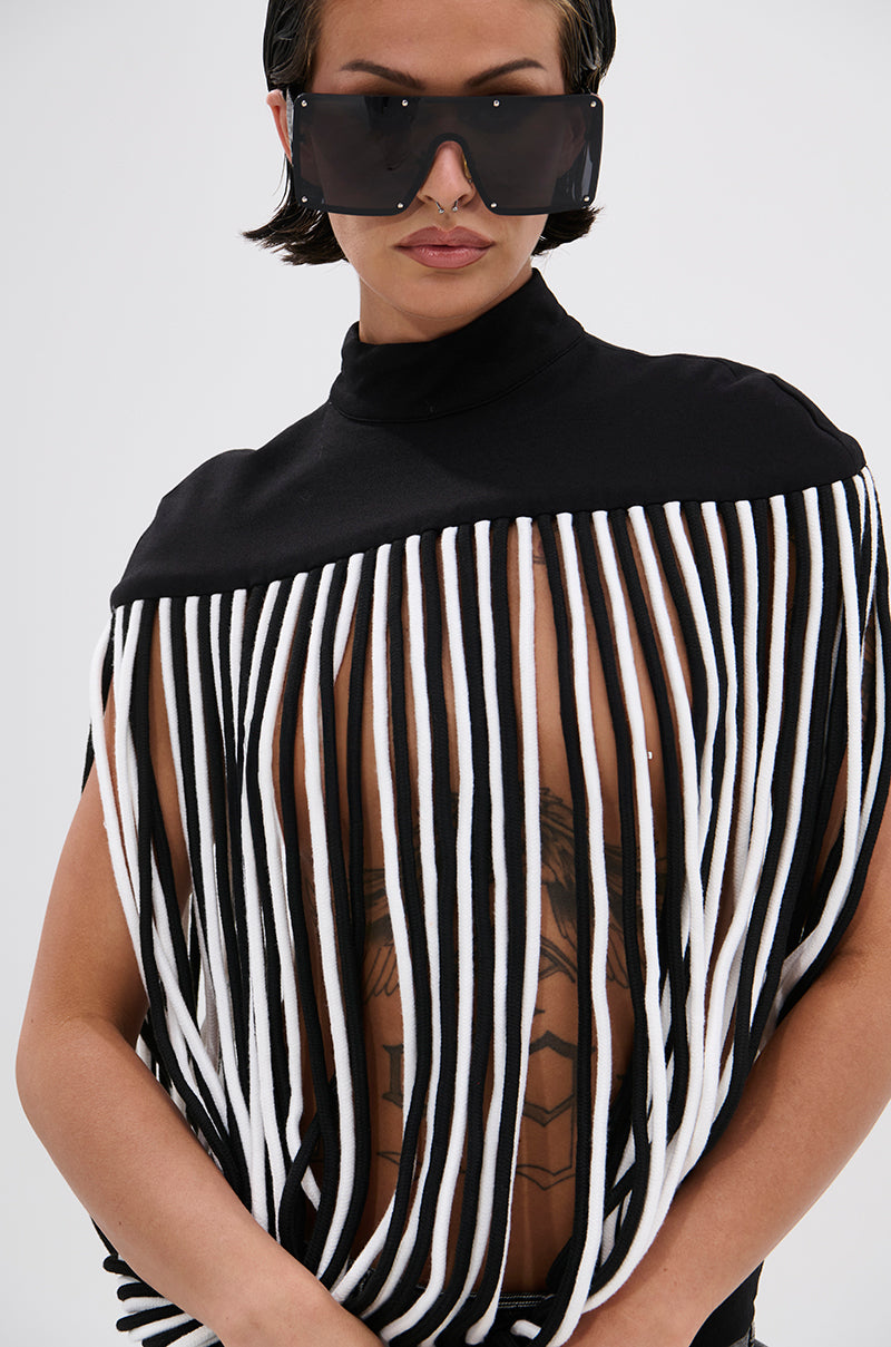 A person in oversized dark sunglasses and a POWER TRIP FRINGE BLOUSE with long black and white fringe and a sculptural mock neckline reveals a geometric chest tattoo, all set against a plain, light background.