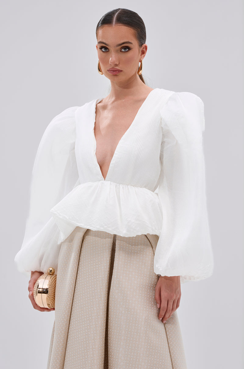 A woman wears the MIMI V-NECK BLOUSE IN WHITE featuring long sleeves, peplum hem, and puff sleeves. She pairs it with beige high-waisted pants, gold hoop earrings, a small gold clutch, and has slicked-back hair.