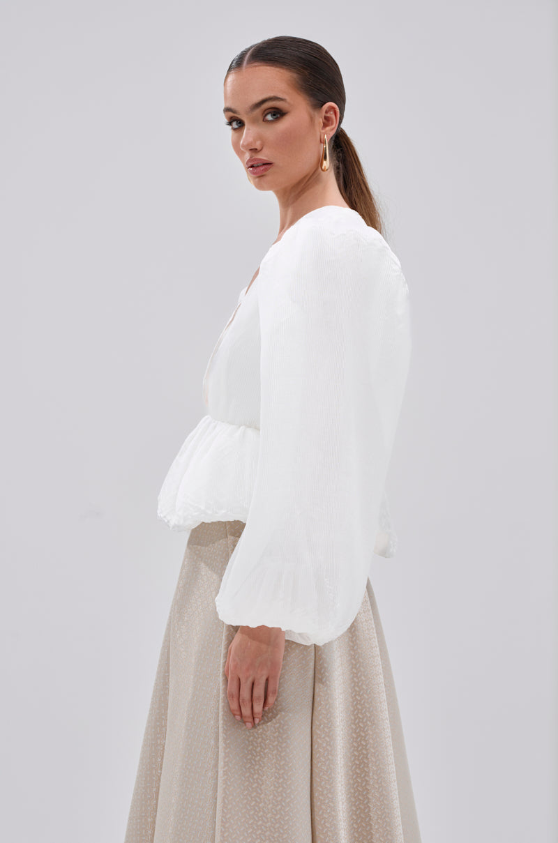 A woman with long brown hair in a low ponytail models the MIMI V-NECK BLOUSE IN WHITE, featuring sheer puff sleeves. She stands sideways, looking over her shoulder against a plain background.