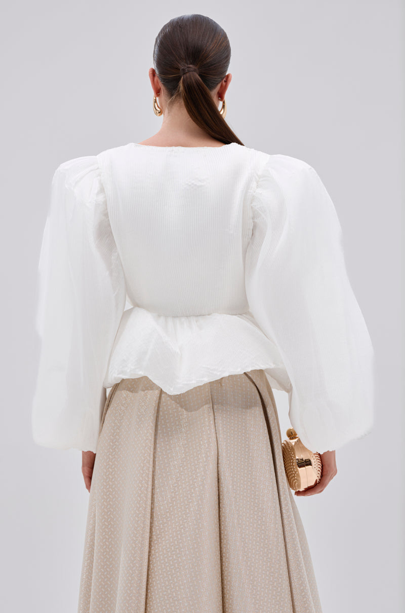 A woman with long, straight brown hair in a low ponytail wears the MIMI V-NECK BLOUSE IN WHITE, paired with a beige pleated skirt and holding a small gold handbag, photographed from behind against a plain background.