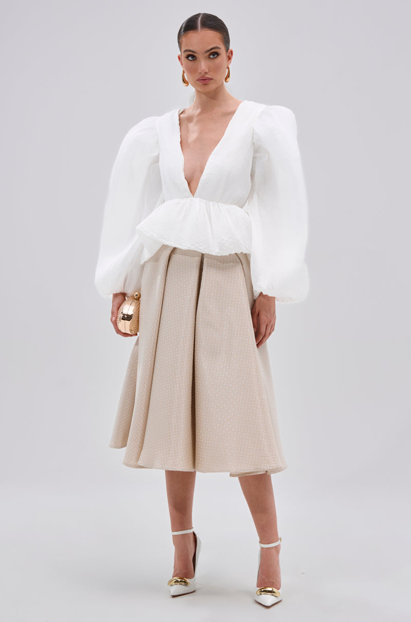 A woman stands holding a small gold clutch, wearing the MIMI V-NECK BLOUSE IN WHITE with puff sleeves, a beige knee-length pleated skirt, white heels with gold tips, and her hair pulled back.