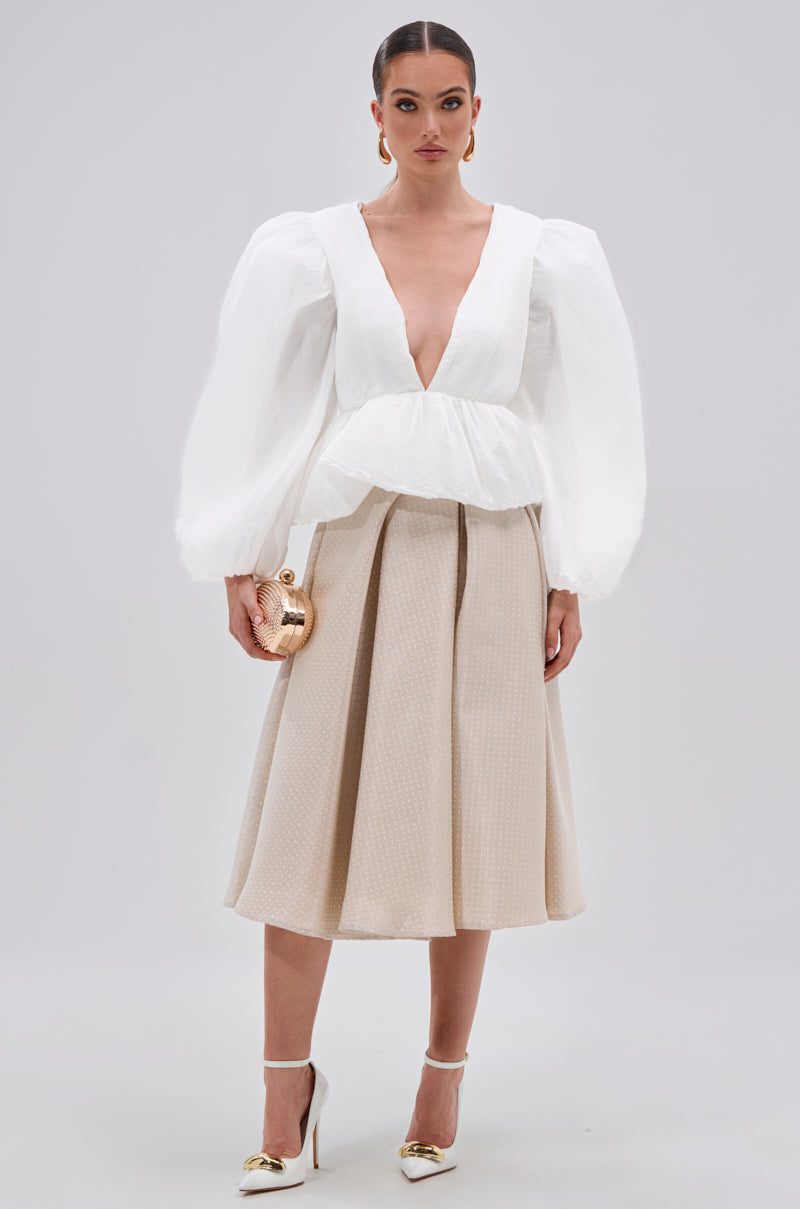 A woman stands against a plain background wearing the MIMI V-NECK BLOUSE IN WHITE with puff sleeves, a beige pleated midi skirt, white heels with gold tips, and holding a small gold purse.