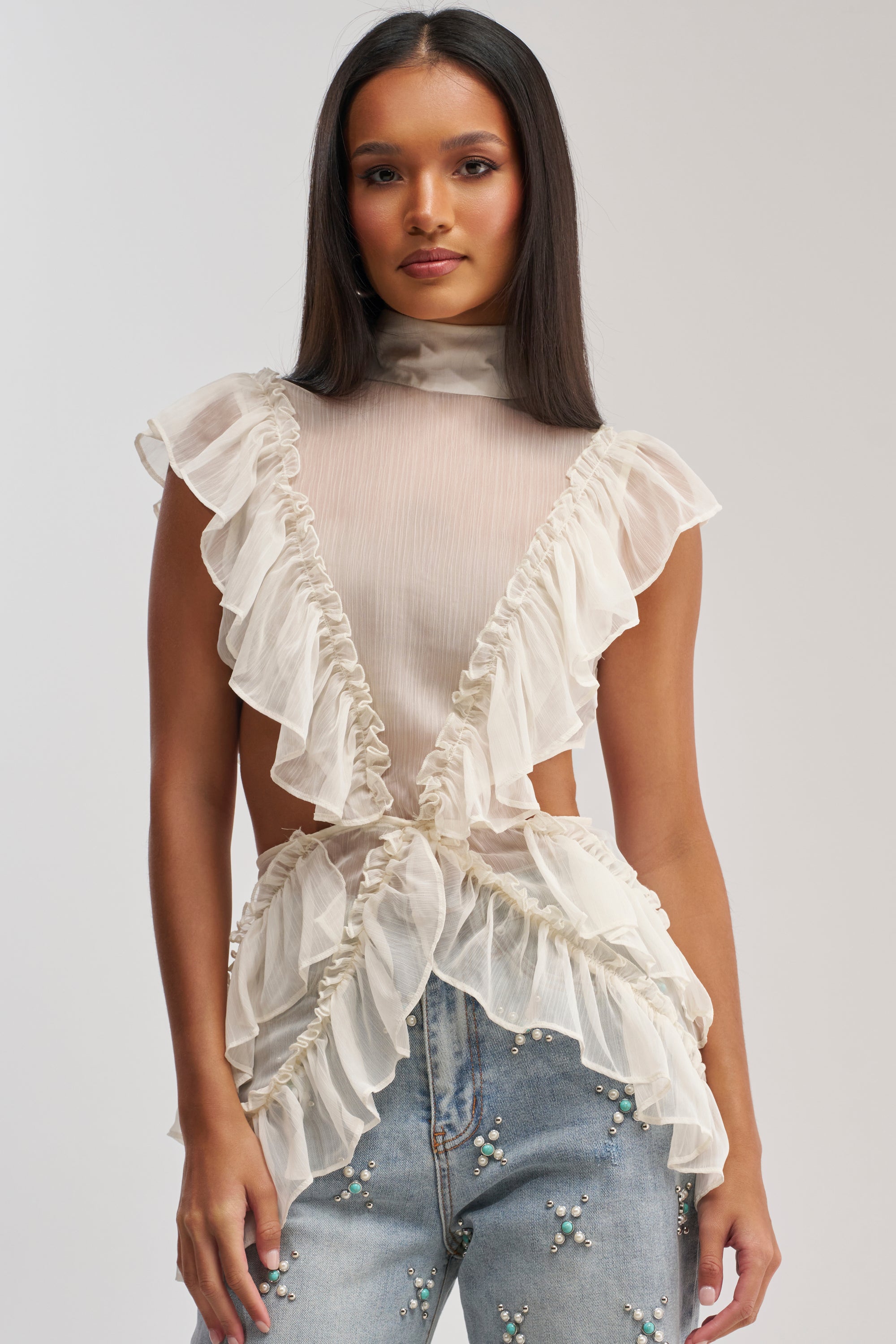 Wearing the DRIFT AWAY BLOUSE—a sheer chiffon ruffle cutout top with open sides—paired with high-waisted, light-wash jeans adorned with pearl-like beads, a woman stands against a plain background.