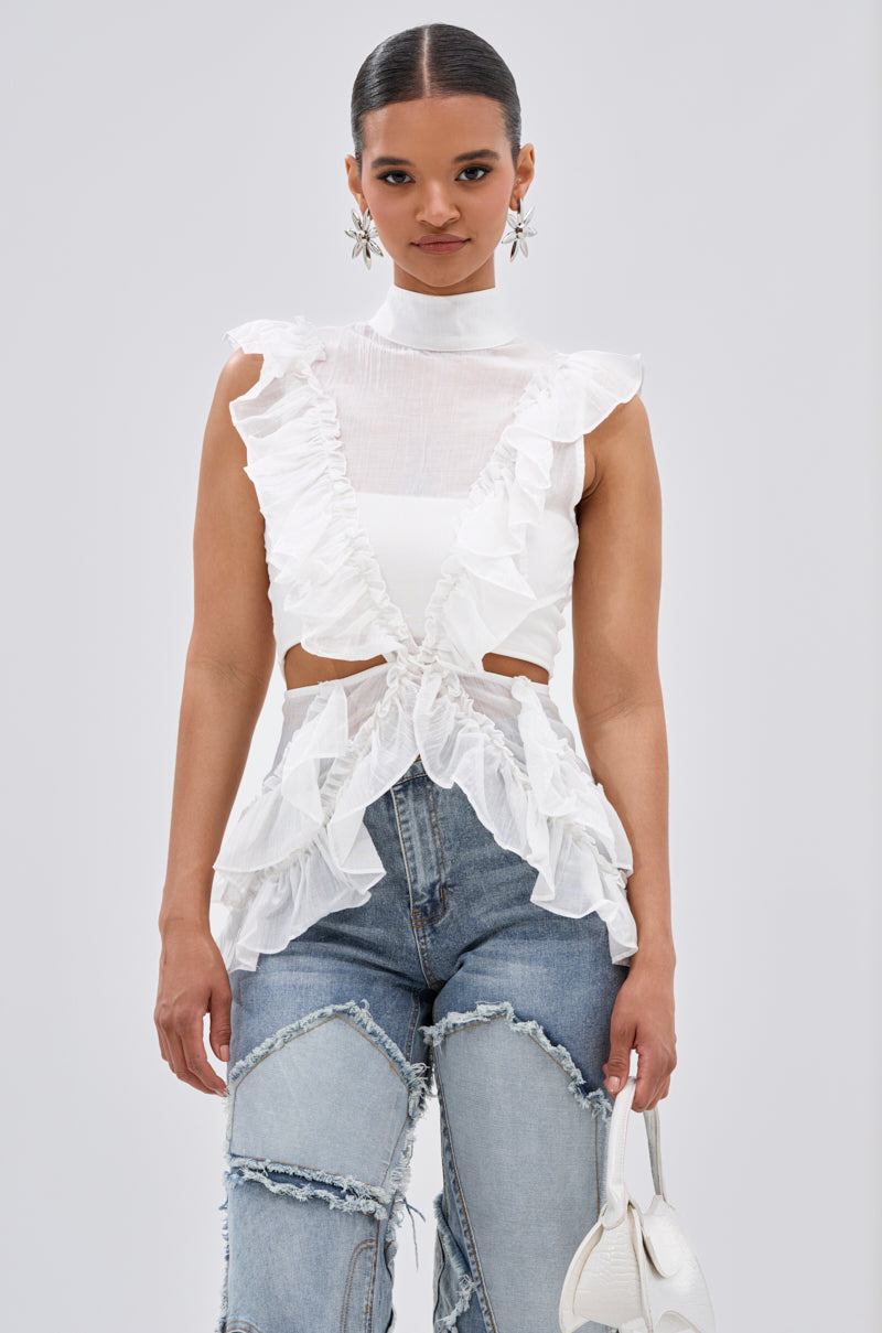 A woman stands against a plain background in the TIFFANY RUFFLE BLOUSE IN WHITE—a sleeveless, ruffled top with cutouts—paired with high-waisted patchwork blue jeans, a small white handbag, statement earrings, and her hair pulled back. 0