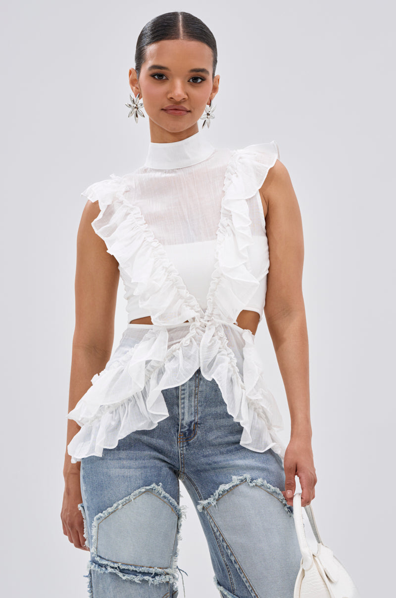 A woman wears the TIFFANY RUFFLE BLOUSE IN WHITE, a sleeveless sheer top with ruffles and a high neck, paired with distressed blue jeans. She carries a white handbag, floral earrings, and has her hair styled back neatly. 1