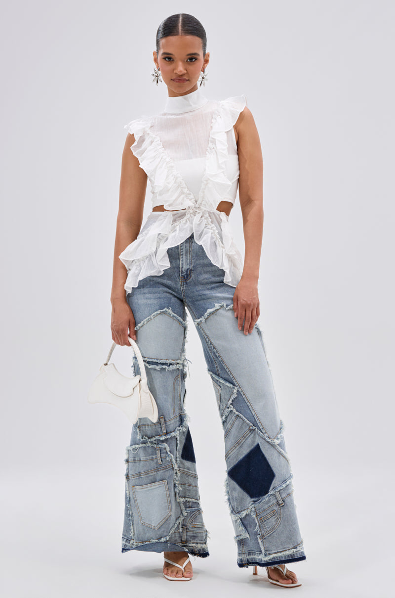 A woman poses against a plain background in the TIFFANY RUFFLE BLOUSE IN WHITE, patchwork wide-leg jeans, white heeled sandals, statement earrings, and holding a small white handbag. 5