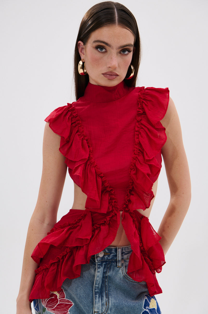 A woman with straight dark hair and gold hoop earrings wears the TIFFANY RUFFLE BLOUSE IN RED, a sleeveless crinkled chiffon top with cut-out sides, paired with floral embroidered blue jeans, standing confidently against a plain background. 0