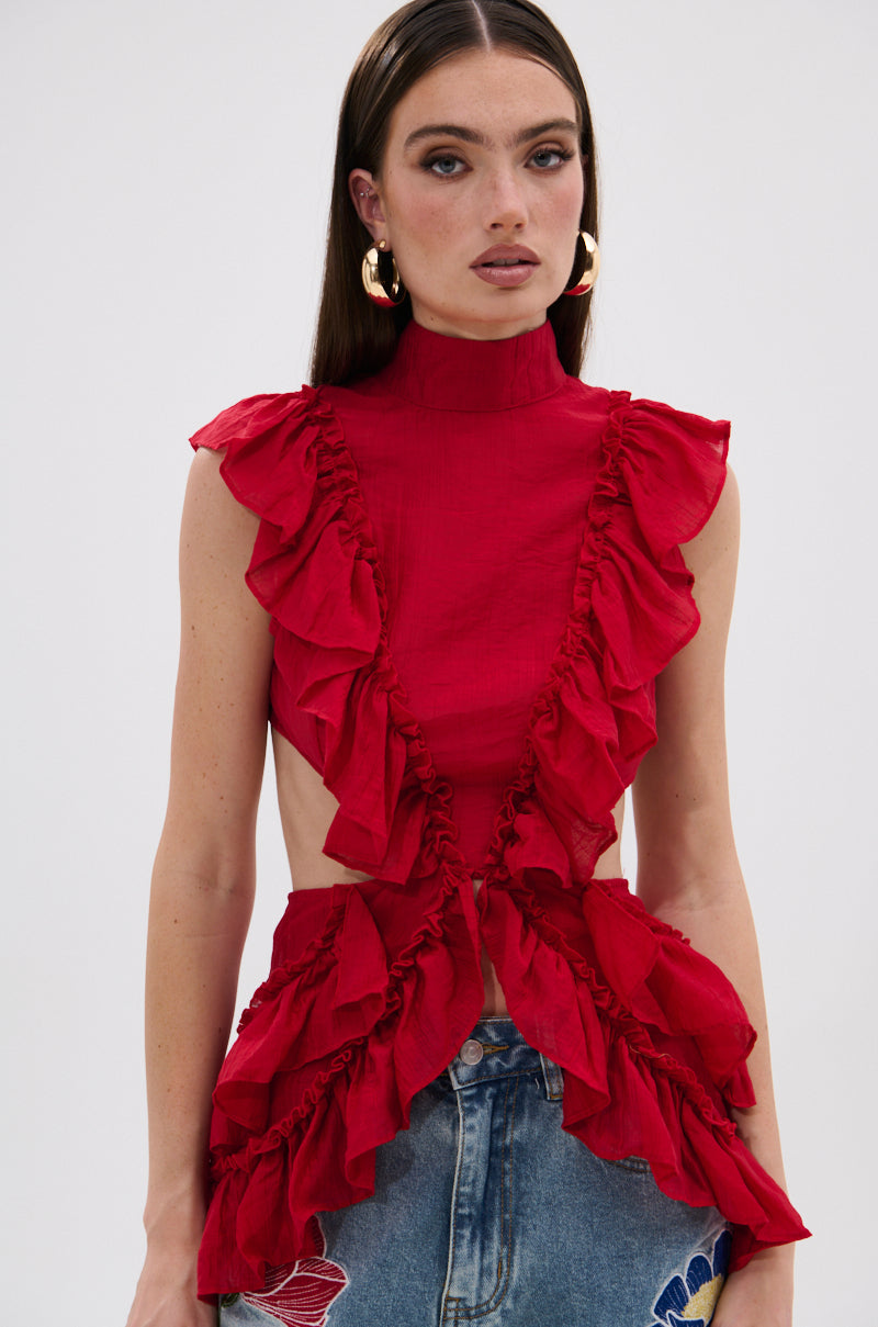 A woman with straight brown hair wears the Tiffany Ruffle Blouse in red, a sleeveless, high-neck style with open sides, paired with blue jeans with embroidered flowers. She completes her look with bold makeup and gold hoop earrings. 1