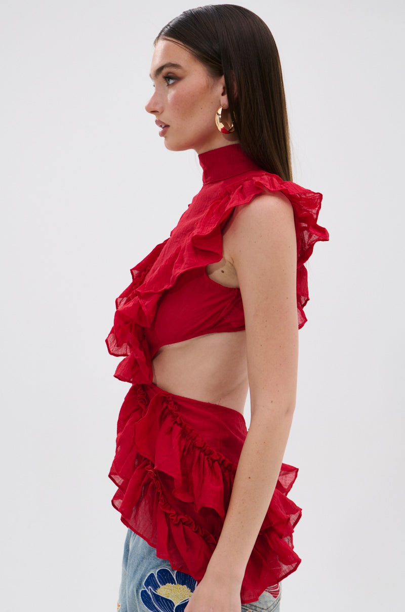 A woman in profile wears the TIFFANY RUFFLE BLOUSE IN RED, made of crinkled chiffon, styled with light blue floral embroidered jeans. She has straight brown hair and gold hoop earrings. 2