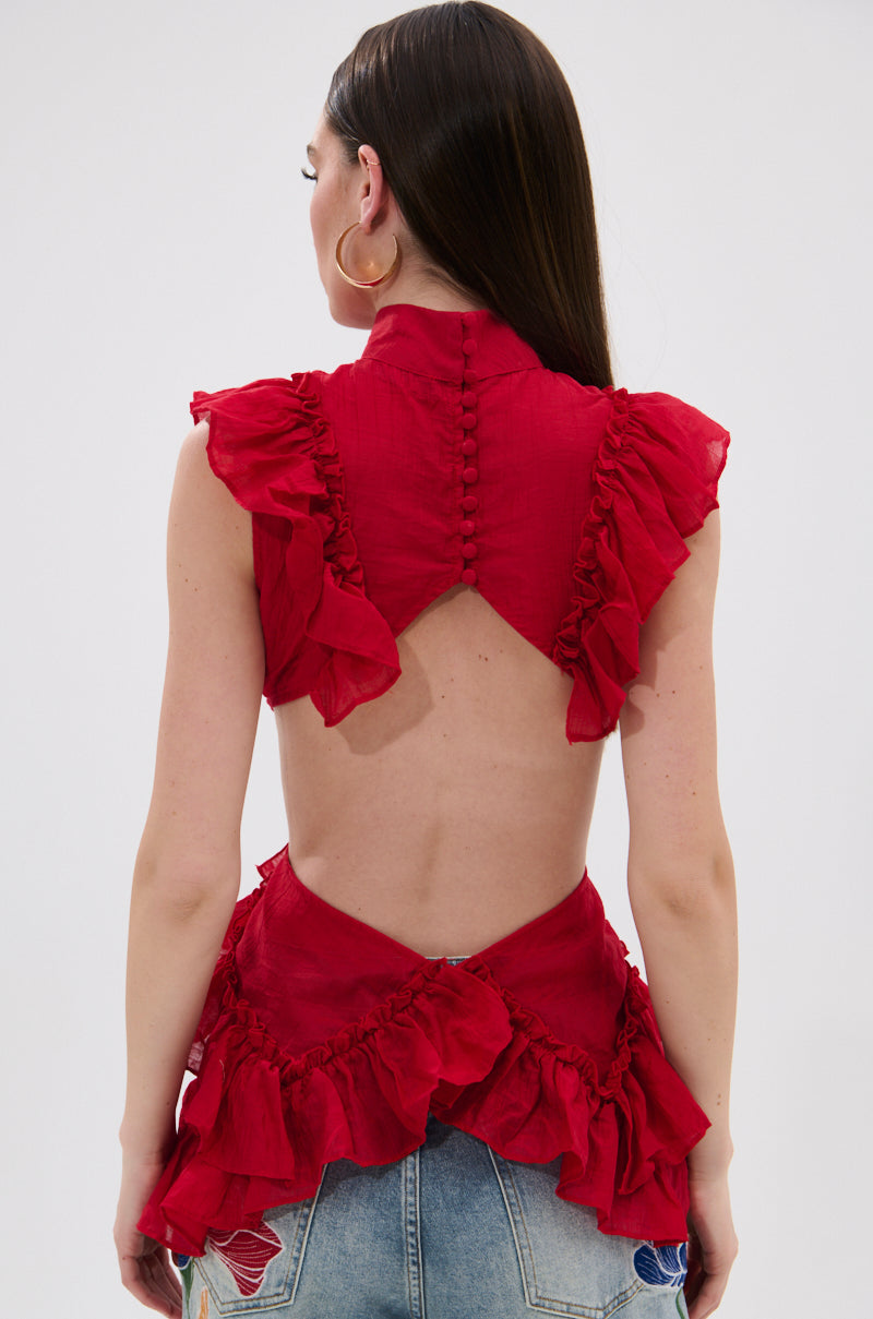 A woman with long brown hair faces away, showing off the open back and layered ruffles of the TIFFANY RUFFLE BLOUSE IN RED—a backless, semi-sheer crinkled chiffon top with buttons—paired with distressed blue jeans. 3
