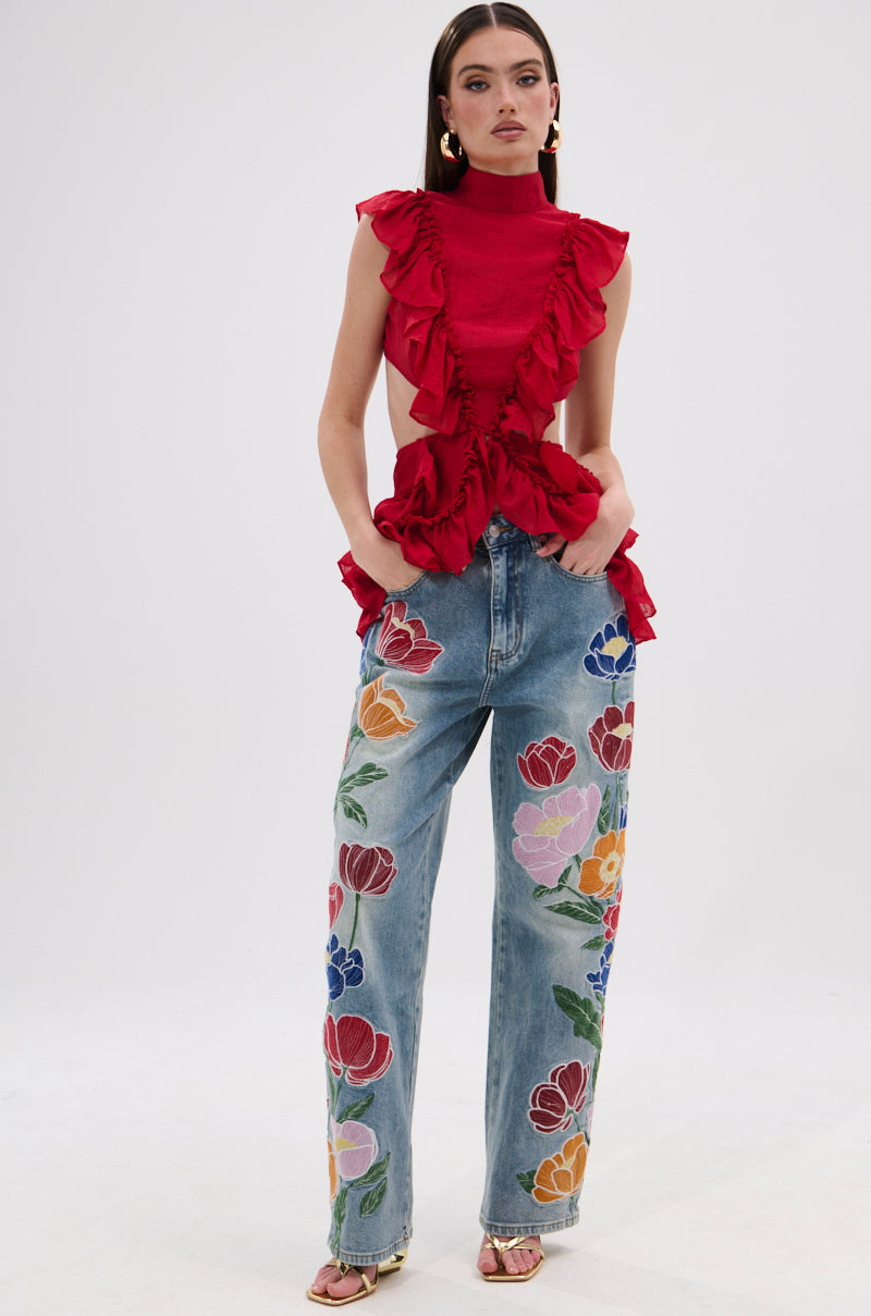 A woman wears the TIFFANY RUFFLE BLOUSE IN RED—a sleeveless, semi-sheer crinkled chiffon top—paired with high-waisted light-wash jeans with colorful floral embroidery, standing against a plain white background. 4