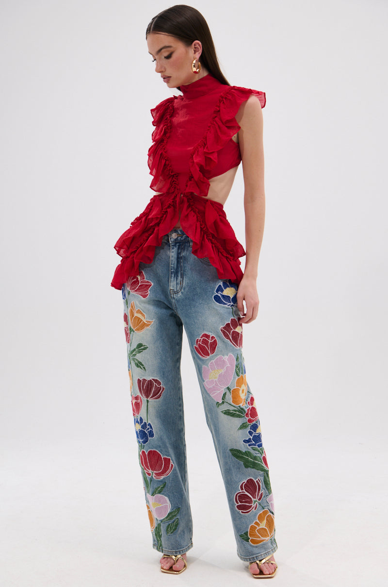 A woman stands confidently against a plain white background wearing the TIFFANY RUFFLE BLOUSE IN RED—a sleeveless, crinkled chiffon top with side cutouts—paired with high-waisted blue jeans featuring colorful floral prints. 5
