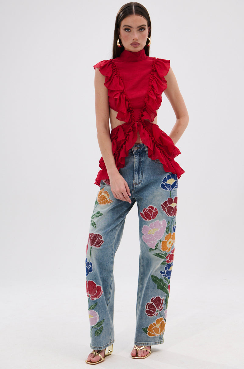 A woman with long, straight hair stands against a plain background in a sleeveless TIFFANY RUFFLE BLOUSE IN RED, crafted from crinkled chiffon, paired with high-waisted blue jeans with colorful floral embroidery, gold sandals, and earrings. 6