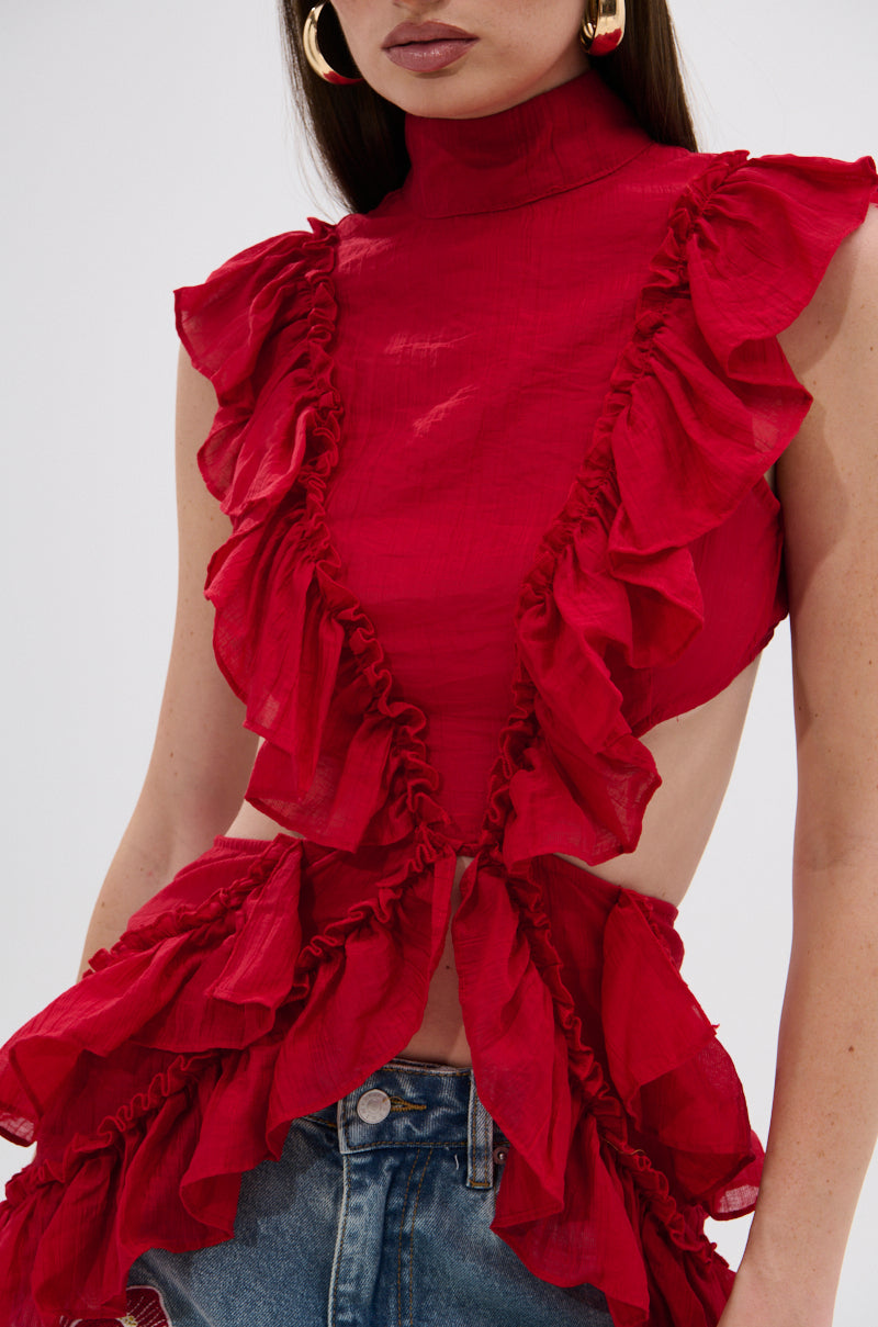 A woman wears the sleeveless, high-neck TIFFANY RUFFLE BLOUSE IN RED with open sides and distressed blue jeans. She adds bold gold hoop earrings and leaves her hair loose to complete the look. 7
