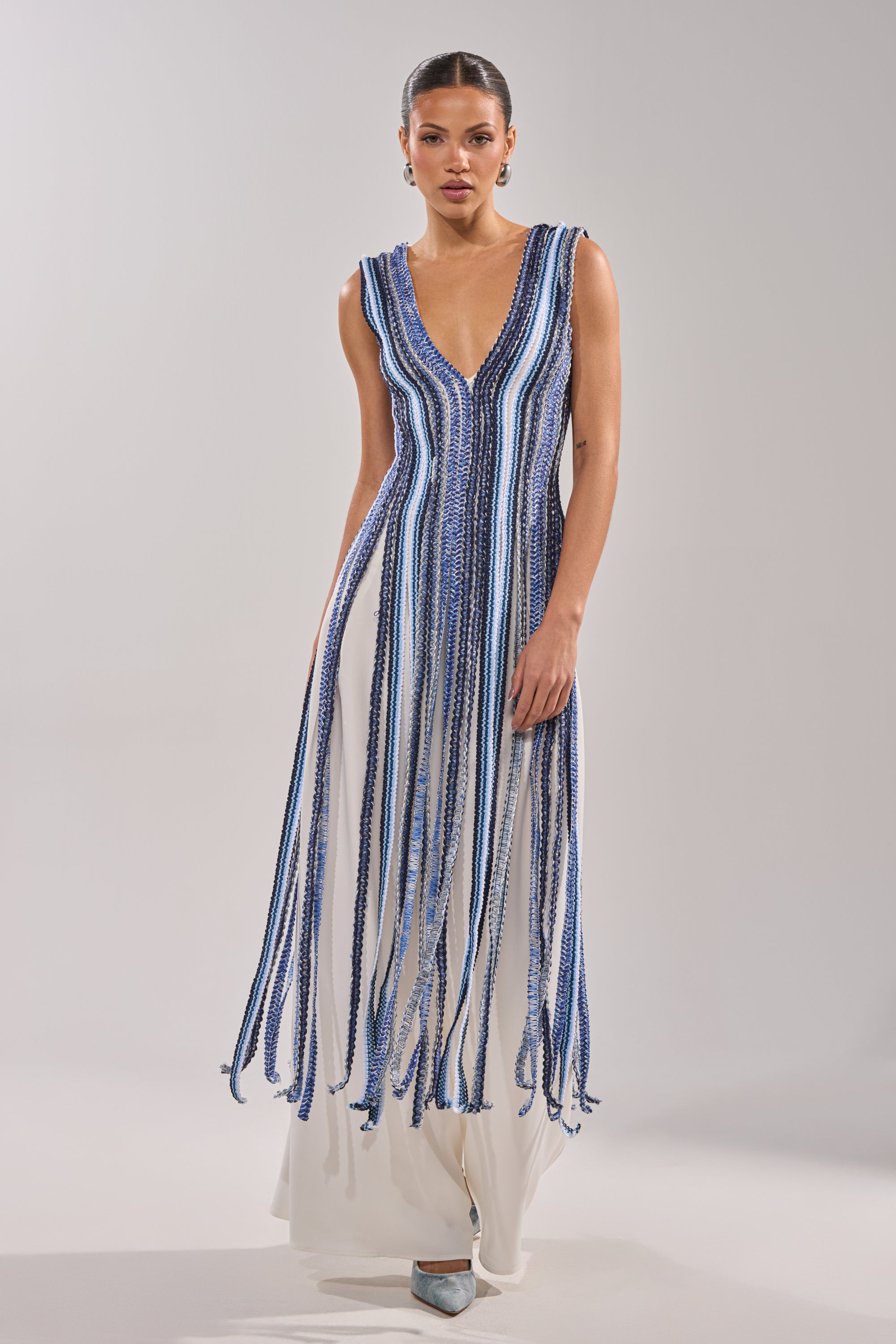 A woman wears the TIED DOWN BRAIDED ROPE TOP—a sleeveless white top with long vertical blue and white fringe and a deep V-neckline—styled with silver heels against a plain light background.
