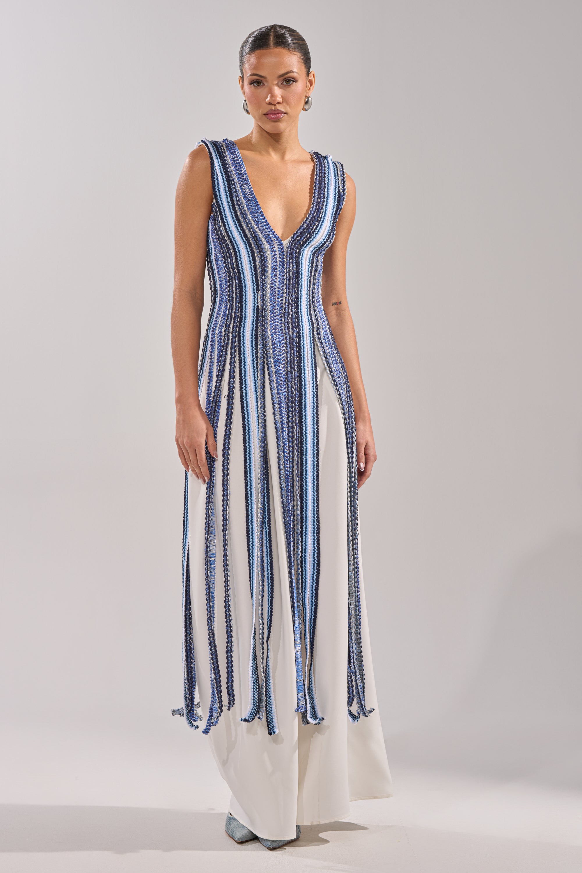 A woman wears the TIED DOWN BRAIDED ROPE TOP, styled resort-ready with vertical blue, white, and black stripes on a white base. She pairs it with wide, flowing pants, has her hair tied back, and accessorizes with earrings.