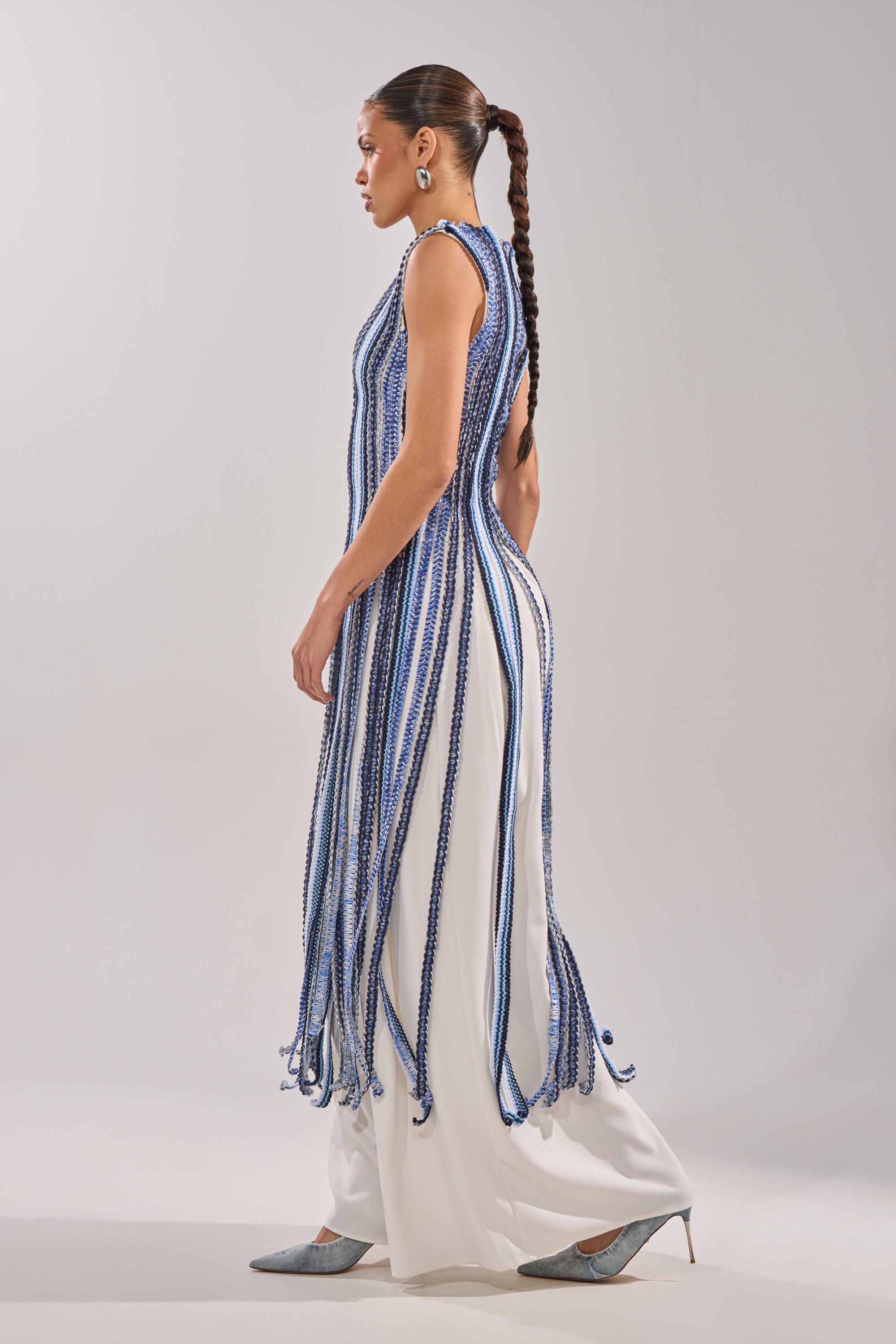 A woman with a long braided ponytail wears the TIED DOWN BRAIDED ROPE TOP—a sleeveless white dress with vertical blue and white strands and braided rope details—paired with silver pointed shoes, standing against a plain light background.