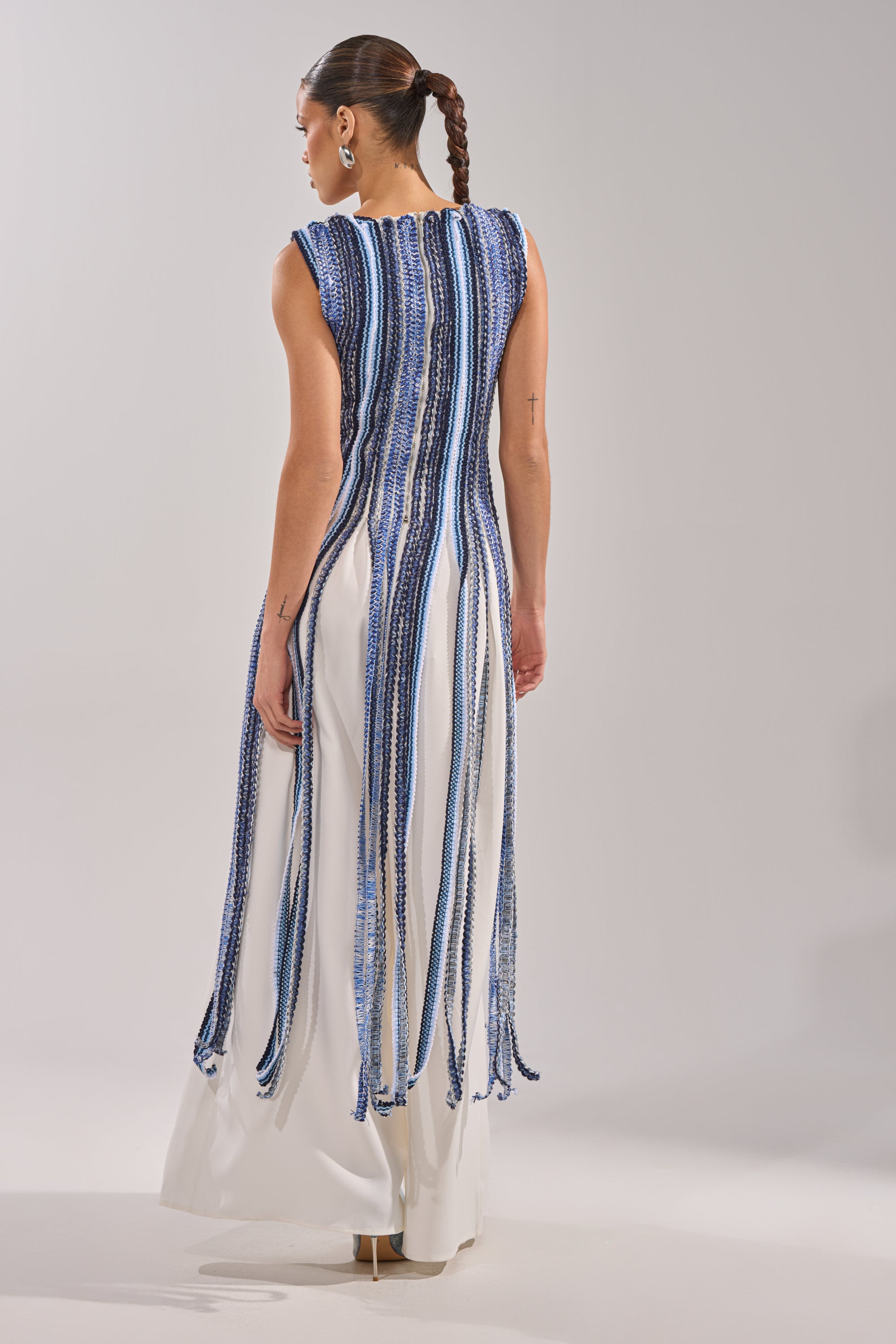 A woman with a braided ponytail, facing away from the camera against a plain light background, wears the TIED DOWN BRAIDED ROPE TOP—a sleeveless white resort piece featuring long, vertical blue beaded fringe strips.