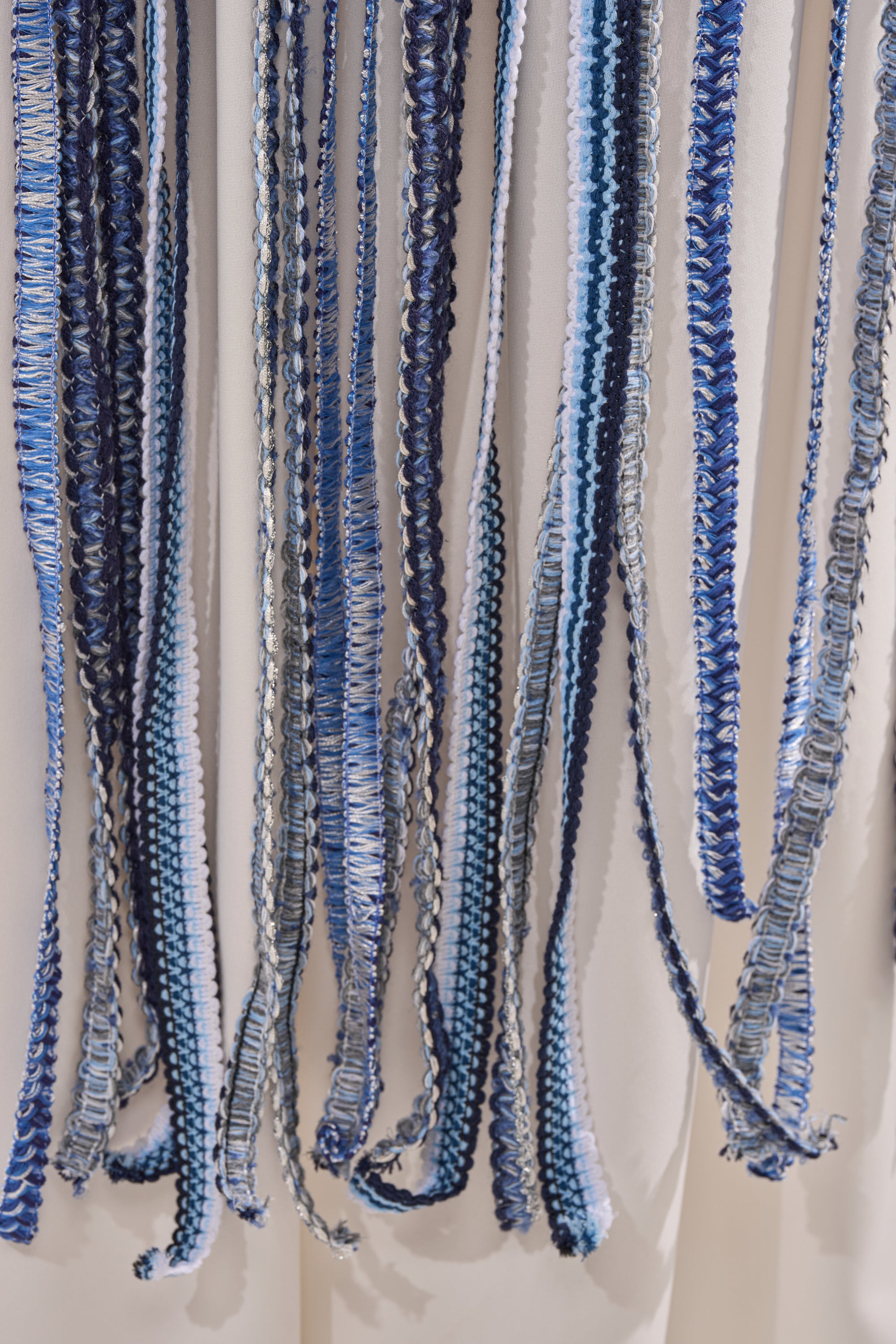 Close-up of the TIED DOWN BRAIDED ROPE TOP featuring multiple strands of blue, navy, and white braided rope cords hanging vertically against a light beige background.