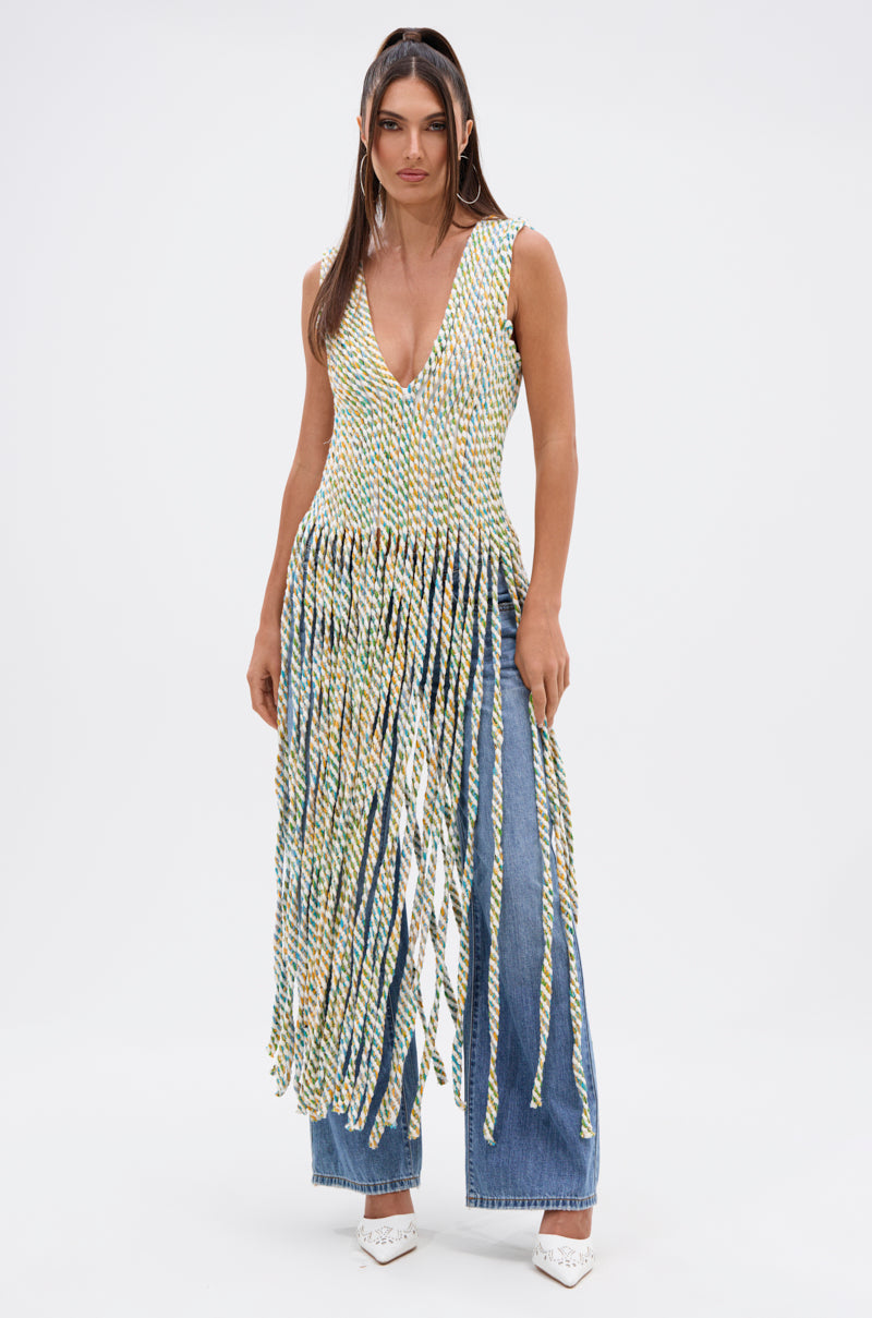 A woman stands against a plain background wearing the TIED DOWN V-NECK ROPE TOP, featuring long multicolored twisted fringe, paired with blue jeans and white heels.
