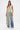 A woman stands against a plain background wearing the TIED DOWN V-NECK ROPE TOP, featuring long multicolored twisted fringe, paired with blue jeans and white heels.
