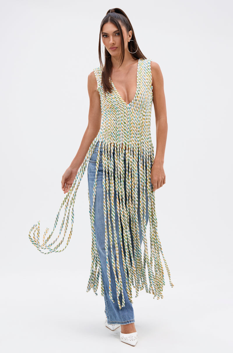 A woman poses against a plain white background in the TIED DOWN V-NECK ROPE TOP, featuring long fringe and multicolored twisted rope details, styled with blue jeans and white heels.