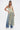 A woman poses in the TIED DOWN V-NECK ROPE TOP, a sleeveless design with colorful twisted cords, styled with wide-leg blue jeans and white pointed heels against a plain white background.