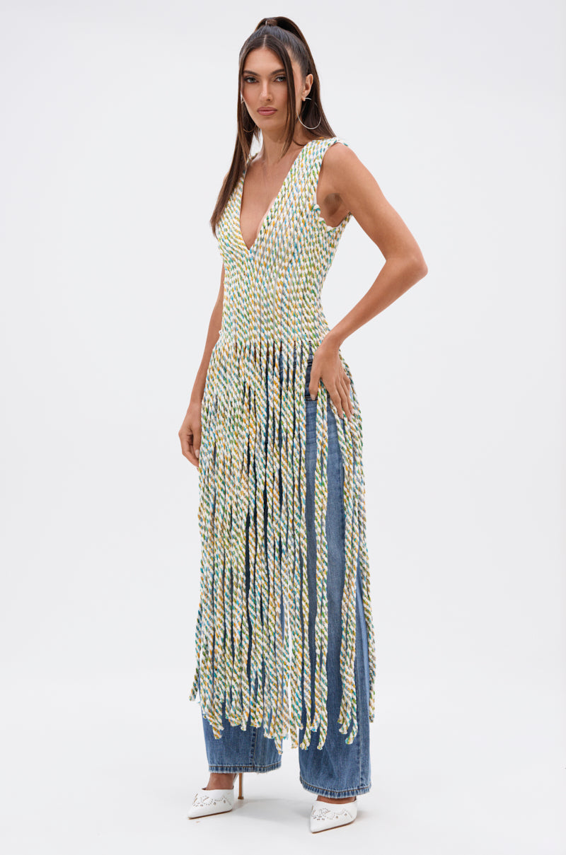 A woman poses against a white background wearing the TIED DOWN V-NECK ROPE TOP, a sleeveless, plunging V-neck multicolored rope top with long fringe, styled over blue jeans and white pointed shoes.