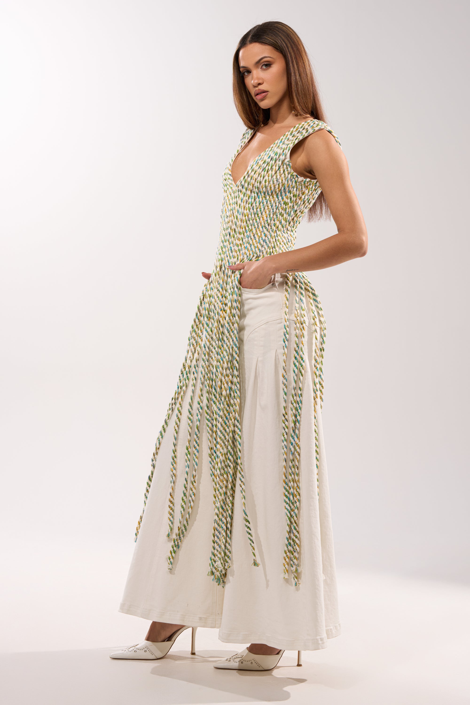 A woman exudes confidence in the TIED DOWN V-NECK ROPE TOP, a sleeveless piece crafted from braided rope, styled with a long white skirt and white heels against a plain light background. 4