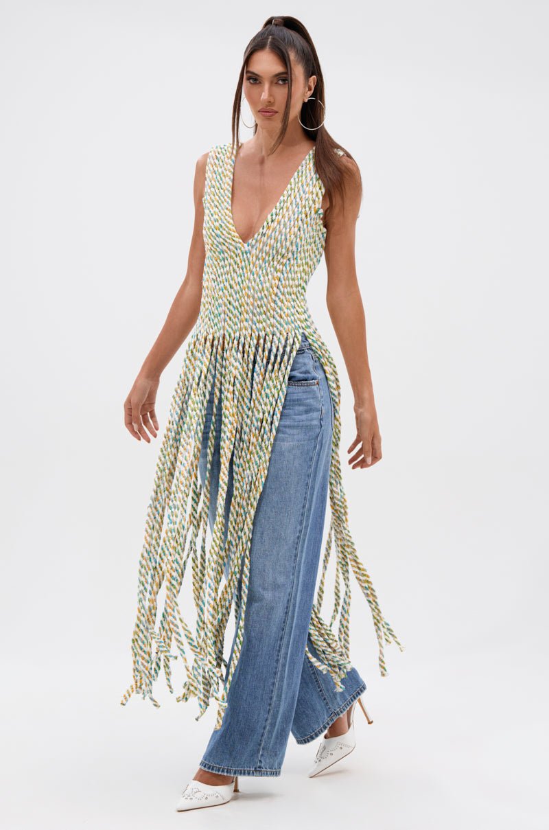 A woman models the TIED DOWN V-NECK ROPE TOP, featuring a sleeveless design and long multicolored twisted fringe, styled with blue wide-leg jeans and white heels against a plain white backdrop.