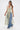 A woman models the TIED DOWN V-NECK ROPE TOP, featuring a sleeveless design and long multicolored twisted fringe, styled with blue wide-leg jeans and white heels against a plain white backdrop.