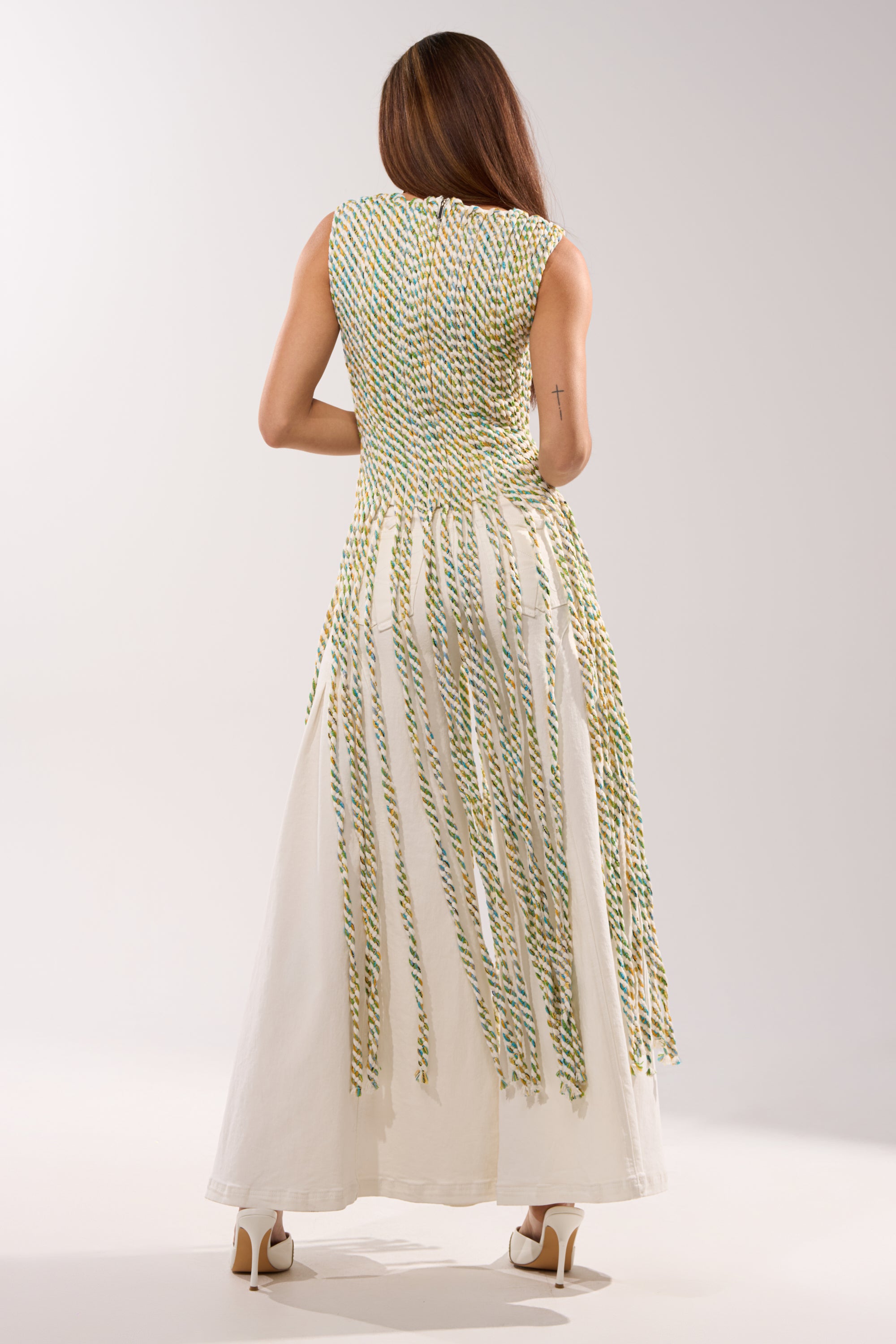 A woman with long brown hair faces away, wearing the TIED DOWN V-NECK ROPE TOP—a sleeveless white dress featuring braided rope details in green, yellow, and blue cascading from the waist—with white high heels. 5