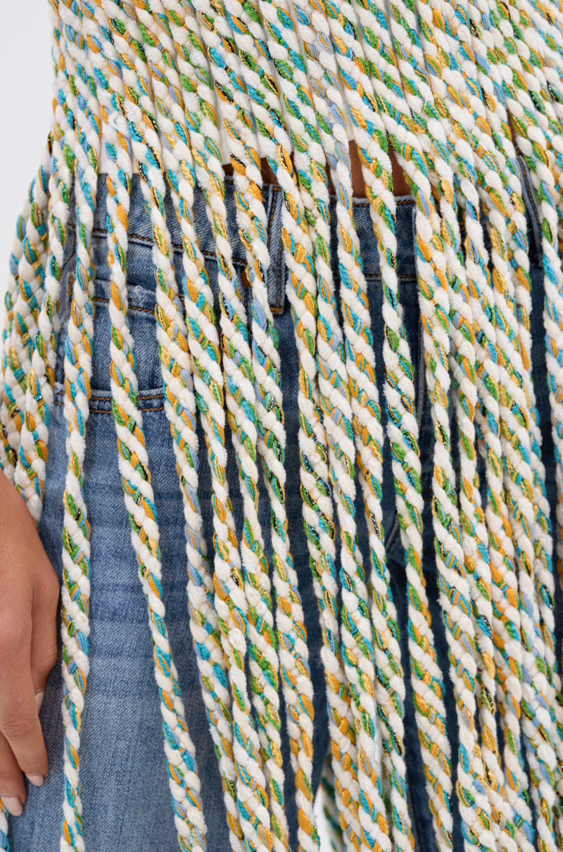 Close-up of a person in blue jeans and the TIED DOWN V-NECK ROPE TOP, a cropped tank with colorful thick twisted rope fringe in white, yellow, blue, and green. The person's hand is partially visible on the left.