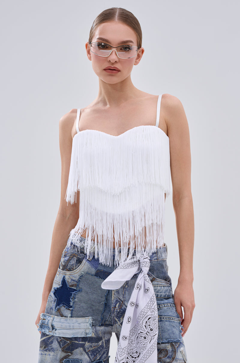 A woman wearing clear glasses, the SHAKE IT FOR ME FRINGE BLOUSE with a metallic zip-back closure, distressed blue jeans with star patches, and a white bandana at the waist stands against a light gray background.
