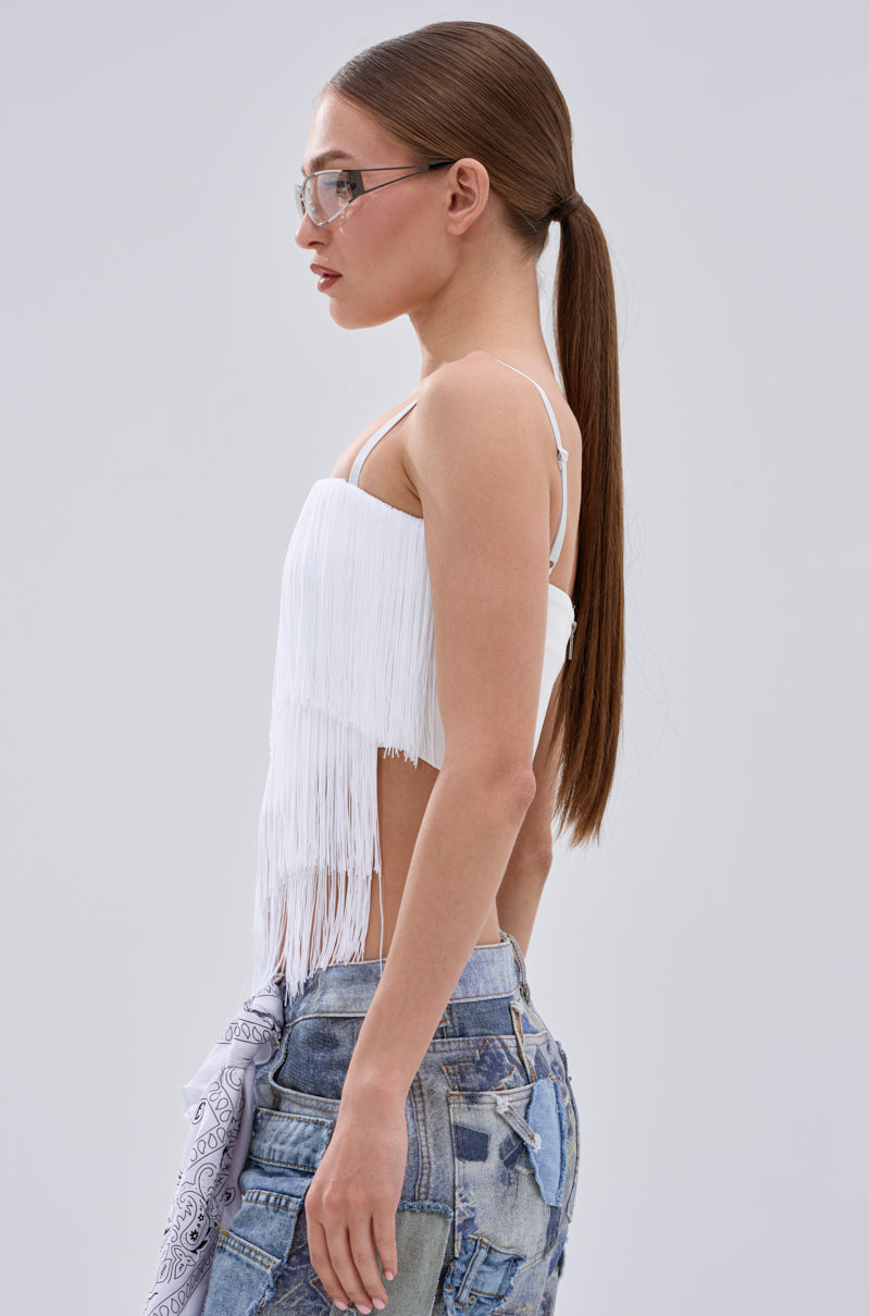 A woman with long, straight brown hair in a ponytail wears glasses and the SHAKE IT FOR ME FRINGE BLOUSE, paired with distressed denim shorts and a light bandana, posing in profile against a plain light gray background.