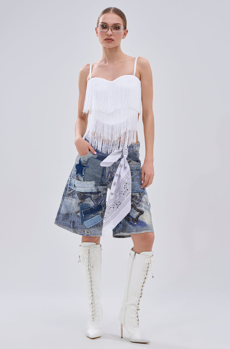 A woman stands against a plain light gray background wearing the SHAKE IT FOR ME FRINGE BLOUSE, a patchwork denim skirt with a white bandana tied at the waist, and knee-high white lace-up boots.