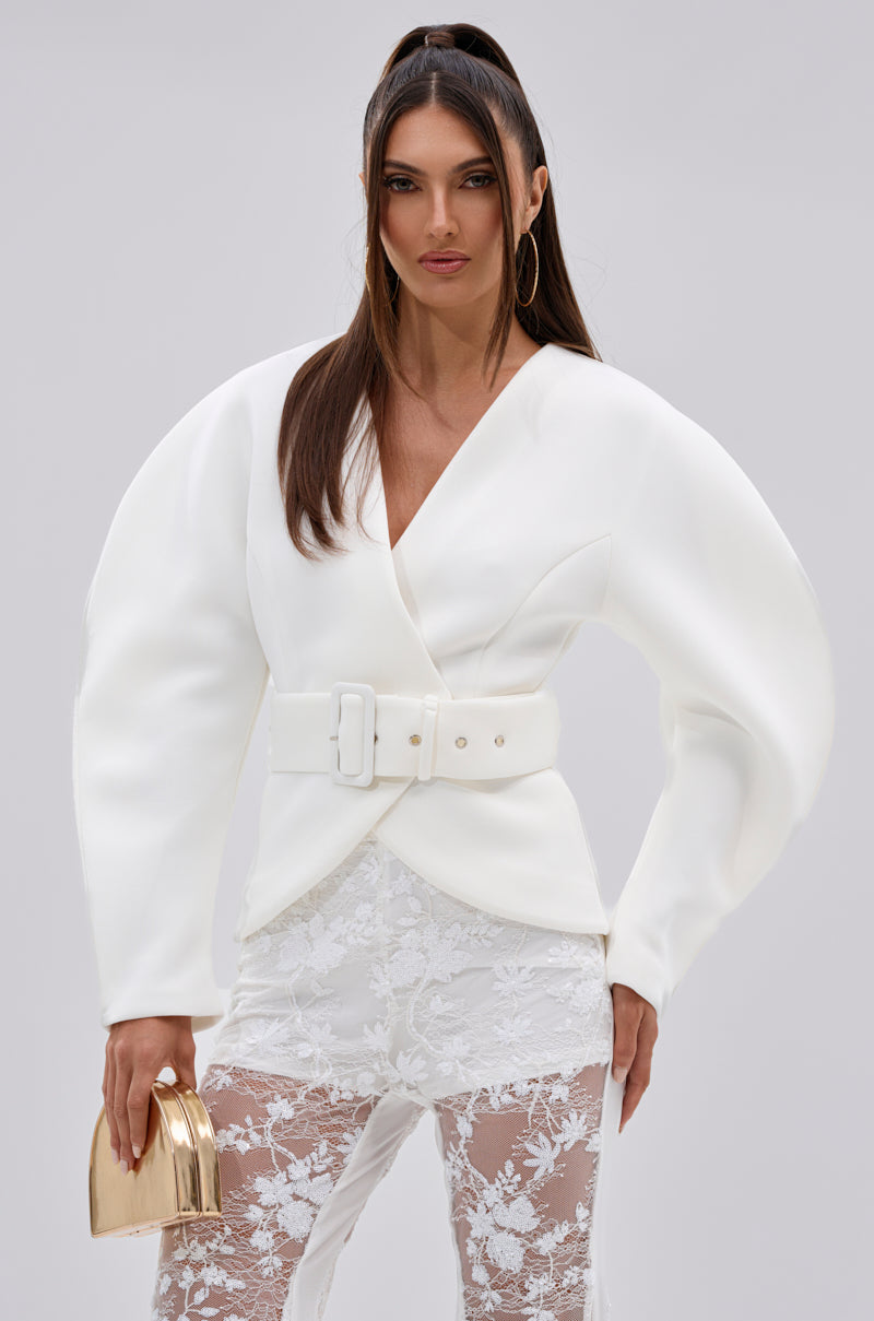 A woman wears the white LANA SCUBA BLOUSE with sculptural sleeves, paired with sheer floral lace pants. She carries a metallic gold mini handbag, sports long half-up hair, and accessorizes with large hoop earrings.